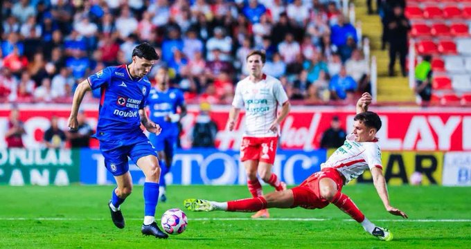 Soccer players in blue and white uniforms compete on a green field during a match, one player in blue controls the ball while another in red slides to tackle, stadium with red seats and crowd in background, sponsor logos visible on jerseys and field.
