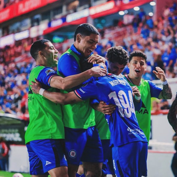 Group of male soccer players in green and blue uniforms stand on green field with stadium seating and crowd in background. Front player wears blue jersey with number 104 and name MICHAL on back facing away. Others in green jerseys hug and gesture excitedly arms raised mouths open. Red and white stadium structure visible behind.