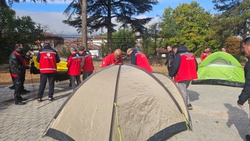 First image shows a large group of people mostly men and some women in red jackets and vests standing in front of a modern building with AFAD sign participants include individuals with beards and short hair posing formally some holding tools. Second image depicts a conference room with long wooden table surrounded by black chairs about 20 people including men and women in red and black clothing seated attentively facing a large screen displaying a landscape image projector visible overhead. Third image features several men in orange and red high-visibility jackets and vests working outdoors under a covered area near trees one man with glasses assisting another in unfolding a large green tarp on the ground white vehicle and excavator in background. Fourth image shows a group of men in red jackets and vests around a beige dome tent on paved ground another green tent nearby trees and fence in background participants appearing to set up or inspect the equipment.
