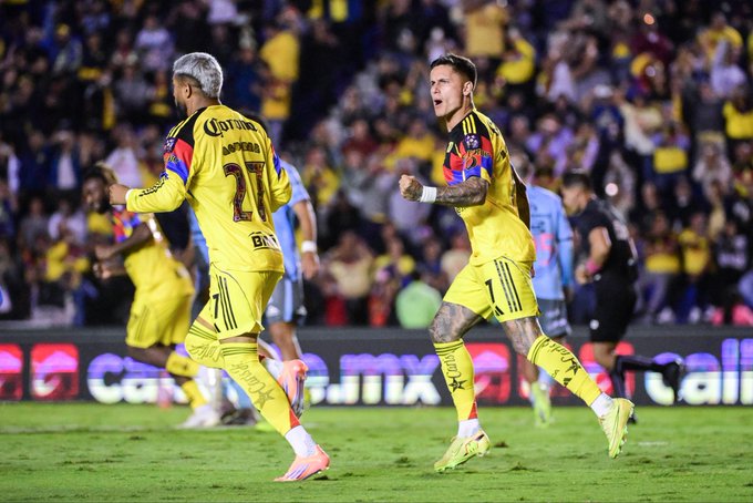Two male soccer players in yellow Club América jerseys with number 27 on one and another number visible celebrate a goal on a green field during a match against Puebla in blue jerseys at Estadio Azulcrema stadium with crowd in background and advertising boards.