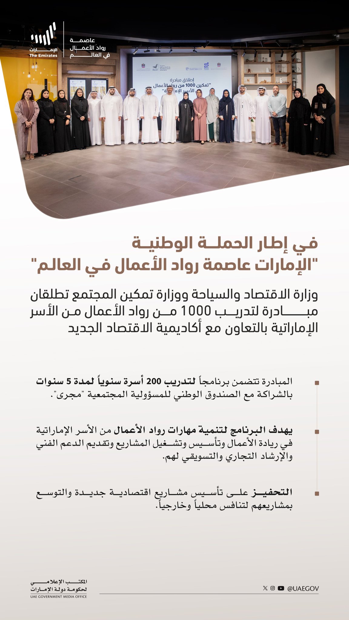 Group of men and women in formal business attire including abayas and suits stand in two rows on a polished floor in a modern indoor hall with yellow walls and a large screen in the background displaying Arabic text about the UAE entrepreneurs training initiative. The individuals appear professional and composed posing for a group photo. Arabic overlay text details the campaign for training 1000 Emirati entrepreneurs by Ministry of Economy and Ministry of Community Empowerment with New Economy Academy.