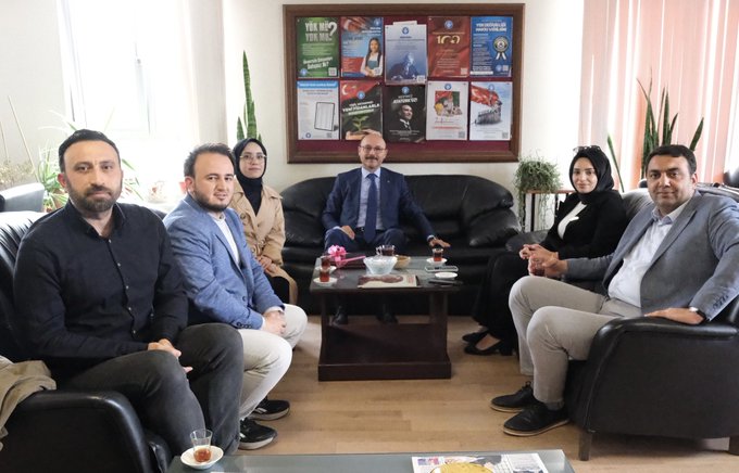 Group of seven professionals seated in a modern office with wooden floors and beige walls. Central table holds glasses of tea and a small vase with flowers. Background features a bulletin board with posters of educational events and organizations. Individuals wear formal attire including suits jackets and headscarves. Plants and curtains add to the setting. Sunlight streams through windows.