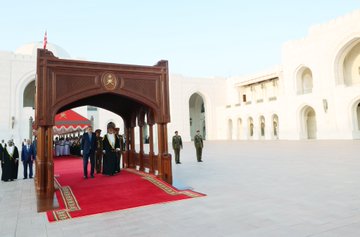 First image shows two men standing side by side in formal attire, one in a dark suit with blue tie identified as Recep Tayyip Erdogan, the other in white robe with gold embroidery and purple headscarf identified as Omani Sultan Haitham bin Tariq, flanked by military personnel in uniforms and hats, against a background with green and red striped canopy and white building. Second image displays a group of people in suits and robes walking on a red carpet under a large wooden archway with intricate carvings and Omani emblem, surrounded by white arched structures and guards in uniforms under a blue sky with Turkish and Omani flags visible. Third image features three men seated in ornate golden armchairs in a lavish hall with green and gold tiled walls and arched doorways, the central figure in white robe and headscarf as Haitham bin Tariq, flanked by suited men including Erdogan in blue tie, with Turkish and Omani flags on stands nearby.