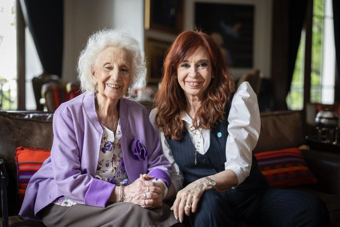 Two elderly women sit closely on a couch in an indoor setting with artwork on walls and large windows. The woman on the left has white hair, wears a purple jacket over a floral blouse, and smiles gently while holding hands. The woman on the right has red wavy hair, wears a white blouse under a vest, smiles warmly, and holds hands reciprocally. Cushions and furniture surround them in a cozy room.