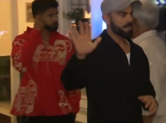 Virat Kohli stands in the foreground wearing a red shirt with white patterns extending his right hand outward in a waving or stopping gesture, his expression neutral with a beard and short hair. Shreyas Iyer is positioned to his right in a black zip-up jacket over a white shirt, looking toward Kohli with a slight smile, both men appearing in their mid-30s with athletic builds. An older man in a white shirt is visible behind them on the left, and the setting includes a beige wall with framed artwork, green potted plants, and soft blue lighting from overhead fixtures, suggesting an indoor restaurant environment.