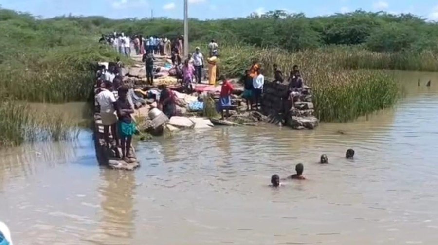 Group of people including men women and children gathered on and around a damaged stone and concrete bridge structure extending over shallow muddy brown water with reeds and vegetation on banks some individuals wading or swimming in water others standing on bridge with belongings like mats and containers in rural setting under clear sky.