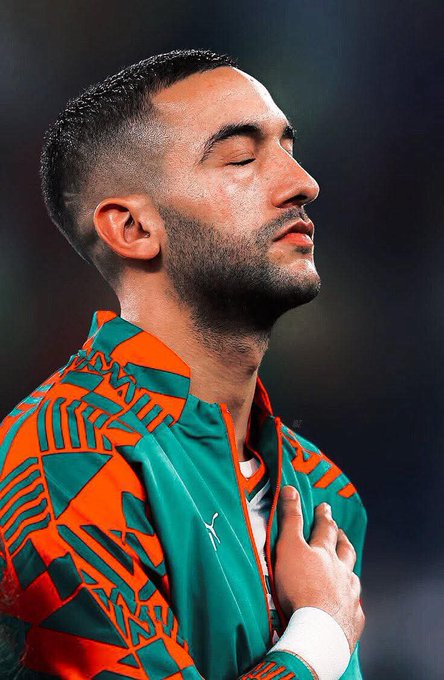 Close-up of a man with short dark hair, beard, closed eyes, and serious expression, wearing an orange and green geometric patterned jacket with white accents, hand placed on chest, against a dark background.