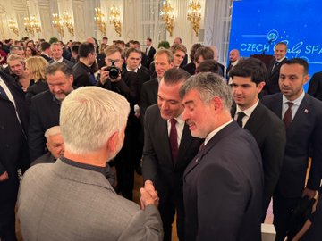 First image shows a group of formally dressed men and women including individuals with gray hair and suits engaging in conversation in a grand room with gold chandeliers and mirrors. Second image depicts three men in suits standing together near a table with a Turkish flag and ornate decor in a elegant room with curtains and a chandelier. Third image captures a crowd of professionals including men with gray and dark hair in suits shaking hands and interacting near a blue screen in a luxurious hall with chandeliers. Fourth image features a group of suited individuals including a man with gray beard holding an open white box containing a clear item while others observe in a opulent room with chandeliers and mirrors.