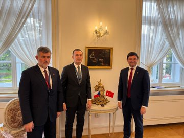 First image shows a group of formally dressed men and women including individuals with gray hair and suits engaging in conversation in a grand room with gold chandeliers and mirrors. Second image depicts three men in suits standing together near a table with a Turkish flag and ornate decor in a elegant room with curtains and a chandelier. Third image captures a crowd of professionals including men with gray and dark hair in suits shaking hands and interacting near a blue screen in a luxurious hall with chandeliers. Fourth image features a group of suited individuals including a man with gray beard holding an open white box containing a clear item while others observe in a opulent room with chandeliers and mirrors.