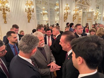 First image shows a group of formally dressed men and women including individuals with gray hair and suits engaging in conversation in a grand room with gold chandeliers and mirrors. Second image depicts three men in suits standing together near a table with a Turkish flag and ornate decor in a elegant room with curtains and a chandelier. Third image captures a crowd of professionals including men with gray and dark hair in suits shaking hands and interacting near a blue screen in a luxurious hall with chandeliers. Fourth image features a group of suited individuals including a man with gray beard holding an open white box containing a clear item while others observe in a opulent room with chandeliers and mirrors.