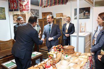 First image shows six professionals in business attire standing around a table with water bottles, boxes, and wooden items at a booth decorated with green plants, patterned fabrics, and informational banners in a modern exhibition hall. Second image depicts several men and a woman in suits shaking hands and conversing near wooden furniture, shelves with decorative boxes, spices, and fabrics under Turkish flag banners in a cultural exhibit space. Third image features a group of people including men and women in formal wear gathered around a table with documents, food trays, and chairs beside a Christmas tree and yellow signage in an indoor venue with red emergency lights.