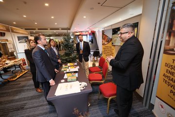 First image shows six professionals in business attire standing around a table with water bottles, boxes, and wooden items at a booth decorated with green plants, patterned fabrics, and informational banners in a modern exhibition hall. Second image depicts several men and a woman in suits shaking hands and conversing near wooden furniture, shelves with decorative boxes, spices, and fabrics under Turkish flag banners in a cultural exhibit space. Third image features a group of people including men and women in formal wear gathered around a table with documents, food trays, and chairs beside a Christmas tree and yellow signage in an indoor venue with red emergency lights.