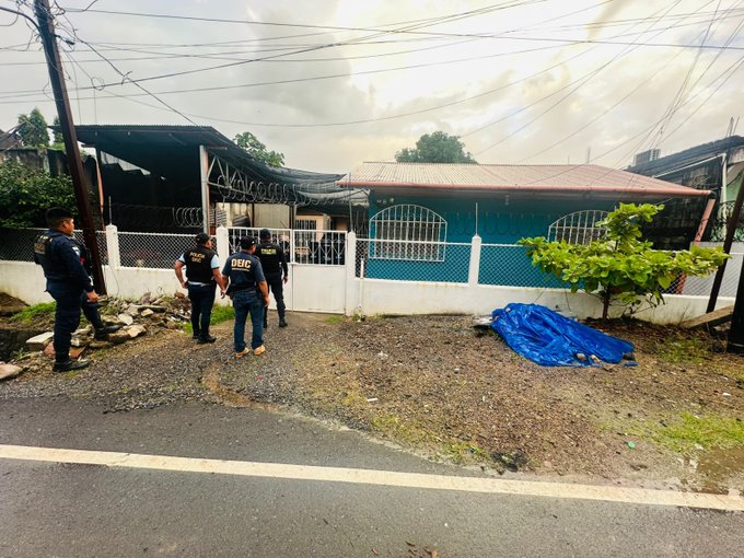 Three police officers in uniforms and vests stand outside a blue house with a red roof surrounded by a white fence and wire mesh in a rural area with power lines overhead a green tarp covering ground nearby and trees visible under overcast skies