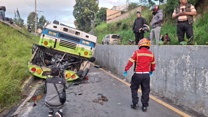 Overturned green and white bus lies on its side on a roadside embankment in a hilly area with grass and trees, emergency responders in red uniforms with helmets attend the scene holding equipment, bystanders in casual clothes observe from nearby vehicles and motorcycles, a photographer kneels capturing the incident, another image shows more firefighters grouped near the damaged bus on the road with additional vehicles and people gathered, third image depicts firefighters in red gear standing along the roadside near the tilted bus with motorcycles and a yellow emergency truck visible in the background under a cloudy sky.