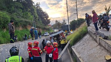 Overturned green and white bus lies on its side on a roadside embankment in a hilly area with grass and trees, emergency responders in red uniforms with helmets attend the scene holding equipment, bystanders in casual clothes observe from nearby vehicles and motorcycles, a photographer kneels capturing the incident, another image shows more firefighters grouped near the damaged bus on the road with additional vehicles and people gathered, third image depicts firefighters in red gear standing along the roadside near the tilted bus with motorcycles and a yellow emergency truck visible in the background under a cloudy sky.