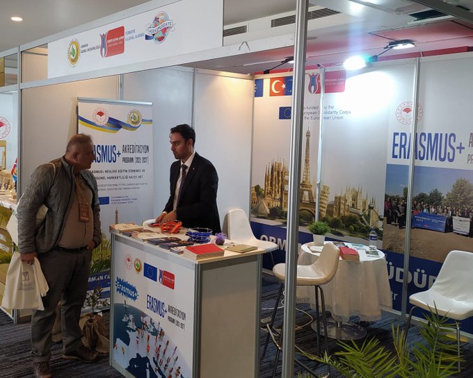 First image displays a group of four professionals standing behind a booth table at an exhibition. The booth features banners with Erasmus+ and ESC logos, Turkish and EU flags, promotional brochures, orange bags, and plants. A man and woman in business attire converse near the setup with Istanbul skyline imagery in the background. Second image shows two men at another booth with materials on Erasmus+ Youth Exchange, chairs, tables with documents, plants, and flags including Turkish and EU ones. The setting includes exhibition hall elements like lighting and neighboring booths.