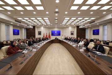 A man in a dark suit with a blue tie and glasses sits at a desk in an office with a Turkish flag on the wall behind him and a screen in the background. Another image shows a long conference table in a modern room with multiple people in formal attire seated around it including women in headscarves discussing with documents water bottles and microphones present and Turkish flags displayed. A third image depicts a similar conference setup with men and women in business suits at a wooden table with laptops papers and flags including a Turkish one in a formal meeting room.