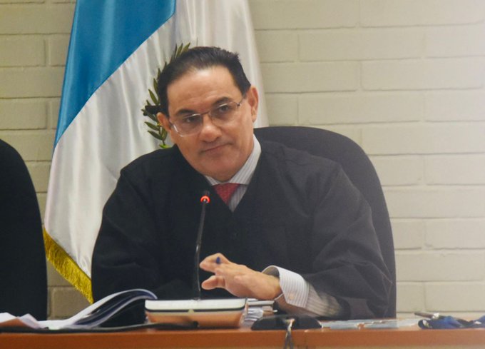 A man identified as Judge Marco Antonio Villeda sits at a wooden desk in a formal room, wearing a black robe, white shirt, red tie, and glasses, with his right hand raised gesturing near a microphone. The Guatemalan flag with blue and white stripes and coat of arms hangs on the wall behind him. Another chair and partial flag are visible on the left, along with papers and a small white object on the desk.