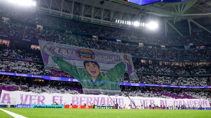 A massive tifo banner hangs in a crowded soccer stadium filled with spectators, depicting a stylized green cartoon character with outstretched arms and an excited expression, accompanied by purple text reading A Ver El Bernabeu Al Competon, with stadium lights illuminating the scene and fans visible in the stands.