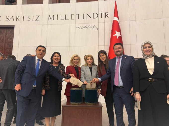 Group of six politicians standing together in formal business attire including suits jackets and headscarves positioned around a wooden table holding two green ballot boxes with papers being placed inside by two women one in red dress and one in gray the background features white marble walls with Turkish flags and inscription Sartsısiz Milletindir in elegant script a wooden door visible on the side all individuals appear professional and engaged in the election process.