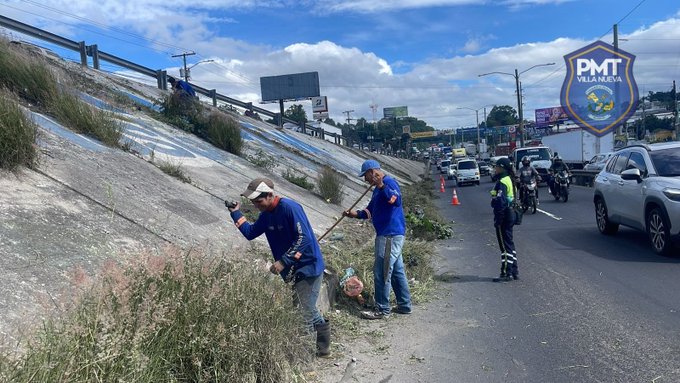 Two uniformed police officers in blue shirts and hats bend down with brooms and tools to sweep and clear dry grass and debris from the edge of a concrete highway embankment. A third officer stands nearby holding a bag. Vehicles including cars and motorcycles pass on the adjacent road under a partly cloudy sky. A large billboard and PMT shield logo are visible in the background. The scene is set along a roadside with chain-link fencing and utility poles.