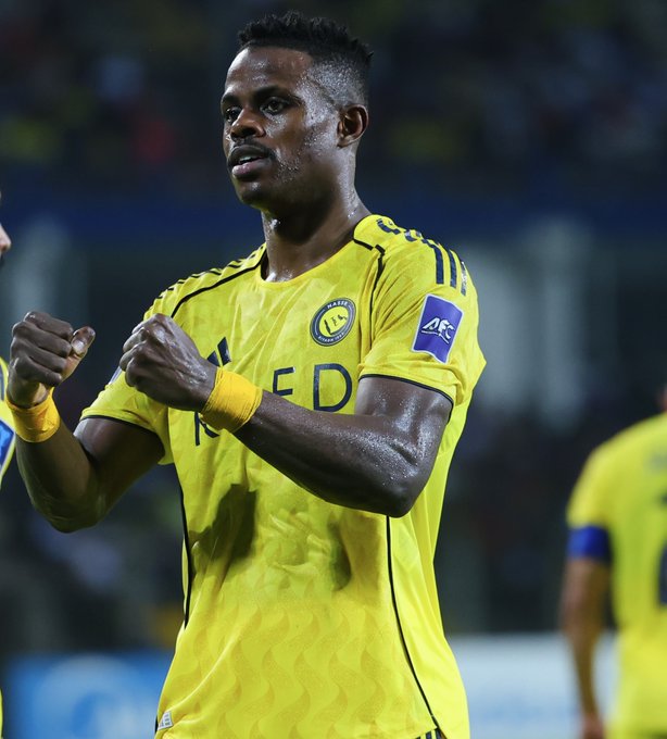 Soccer player Haroun Kamara in yellow Al Nassr uniform with black accents and sponsor patches stands on stadium field raising both fists in celebration surrounded by blurred stadium seating and yellow lighting.