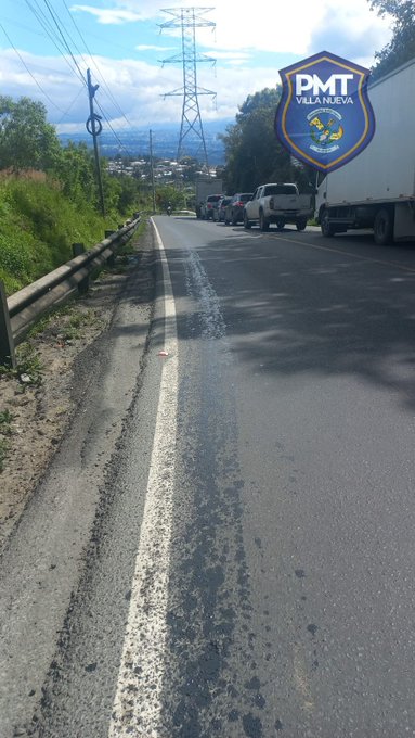 Roadway with white lane markings and oil spill visible on asphalt surface surrounded by green vegetation and roadside barriers several vehicles including a white truck parked nearby under cloudy sky with power lines and transmission towers in background police municipal transit vehicle with blue shield emblem displaying PMT Villa Nueva text positioned prominently on road shoulder