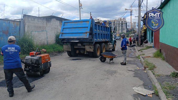 Workers in blue uniforms push wheelbarrows filled with materials toward a large blue dump truck parked on a paved street. One worker operates a small orange compactor machine nearby. The scene includes concrete walls, grass patches, power lines, and a green building with a P.M.T. VILLA NUEVA police badge sign. Construction cranes and apartment buildings appear in the background under a cloudy sky.