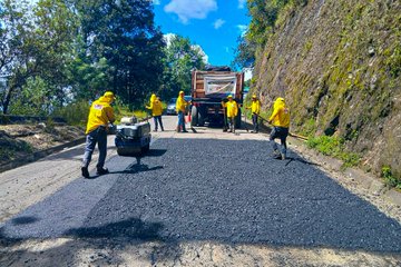 First image shows four workers in yellow raincoats and hats using tools to repair a damaged road section near a white truck and orange cones on a roadside with a rocky mossy cliff and vegetation. Second image displays a white dump truck parked on a paved road beside a cliff with trees and blue sky, with another vehicle in the background. Third image depicts workers in yellow gear spreading fresh black asphalt from a dump truck using rakes on the road surface adjacent to a green hillside and trees under a blue sky. Fourth image illustrates multiple workers in yellow outfits compacting newly laid asphalt with a roller machine near a truck on a road flanked by trees and a cliff.