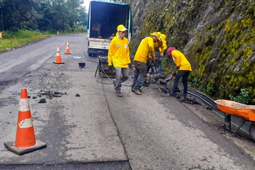 First image shows four workers in yellow raincoats and hats using tools to repair a damaged road section near a white truck and orange cones on a roadside with a rocky mossy cliff and vegetation. Second image displays a white dump truck parked on a paved road beside a cliff with trees and blue sky, with another vehicle in the background. Third image depicts workers in yellow gear spreading fresh black asphalt from a dump truck using rakes on the road surface adjacent to a green hillside and trees under a blue sky. Fourth image illustrates multiple workers in yellow outfits compacting newly laid asphalt with a roller machine near a truck on a road flanked by trees and a cliff.