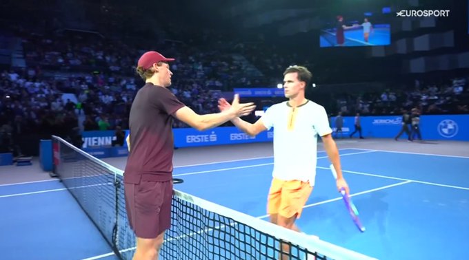 Two male tennis players, Jannik Sinner in a maroon shirt and cap shaking hands with Daniel Altmaier in a white shirt and orange shorts over a tennis net on a blue court surface, with a crowd in the background and an Eurosport screen visible showing the match.
