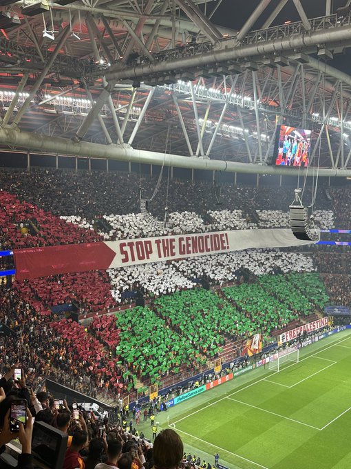 Stadium interior at night with large crowd in stands holding red white and green umbrellas and banners forming the Palestinian flag colors, a prominent horizontal banner with red background and white text reading STOP THE GENOCIDE in bold letters, overhead screens and field visible below, fans in orange clothing on the pitch side.
