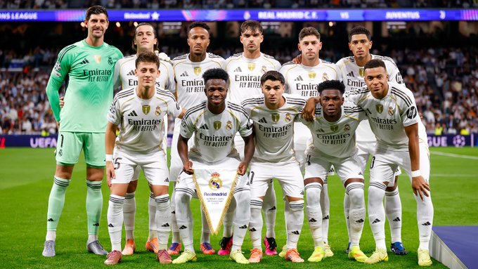 Eleven soccer players from Real Madrid stand in a line on a green field with a stadium crowd behind them. They wear white jerseys with black and purple details and sponsor logos, green shorts and socks for most, except the goalkeeper in green uniform. The group poses with arms around each other shoulders, some smiling, holding a white banner with trophy emblem. UEFA logo visible on scoreboard above.