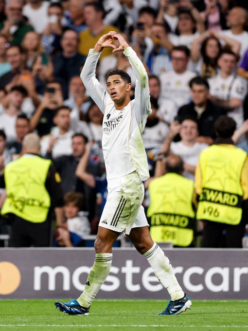 Real Madrid player with curly hair stands on green soccer field raising both arms overhead in celebratory gesture, dressed in white long-sleeve jersey with Adidas stripes and sponsor patches, white shorts, white socks, and blue cleats. Background shows packed stadium stands filled with fans holding white flags and scarves, some taking photos with phones. Yellow-vested security personnel stand nearby on the field edge. Prominent advertising boards display Mastercard logo and other sponsors along the pitch perimeter.
