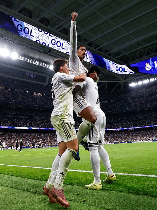 Three Real Madrid players in white uniforms celebrate a goal on a stadium field at night under bright lights with a large blue GOL banner in the background showing raised arms and excited poses amid a crowd of spectators.