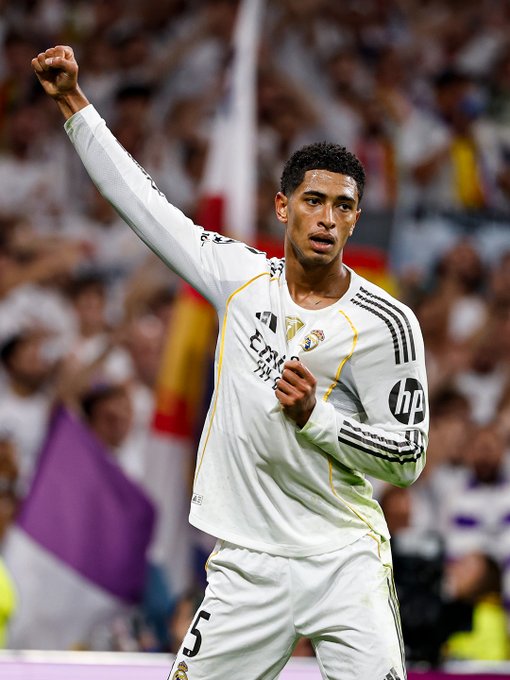 Jude Bellingham raises his right fist in celebration on a soccer field, wearing a white long-sleeve Real Madrid jersey with yellow accents, number 5 on the back, HP sponsor logo, and club emblem, black shorts, and white socks, standing on green turf with stadium seating filled with blurred spectators holding purple and white flags under bright arena lights.