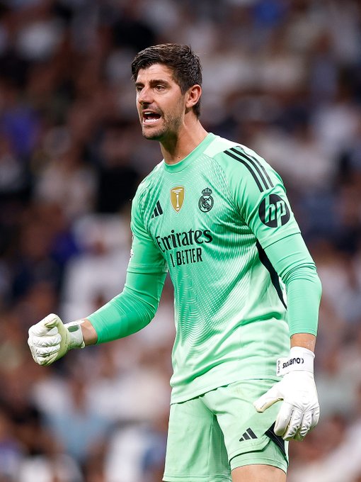 Thibaut Courtois stands as goalkeeper in green Real Madrid uniform with sponsor logos including Emirates and HP on sleeves extending arms with white gloves extended forward in diving motion on soccer field with blurred stadium crowd and green pitch visible behind him.