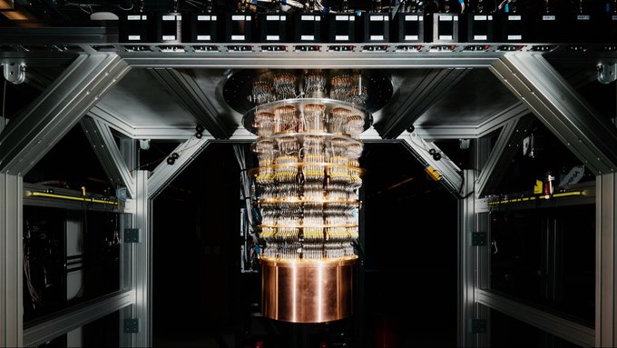 A metallic frame structure houses a tall cylindrical quantum computer apparatus with multiple stacked circular components connected by wires and tubes, featuring a central copper-colored cylinder and surrounding glass-like or transparent elements suspended in a dark enclosure with horizontal support beams and panels.