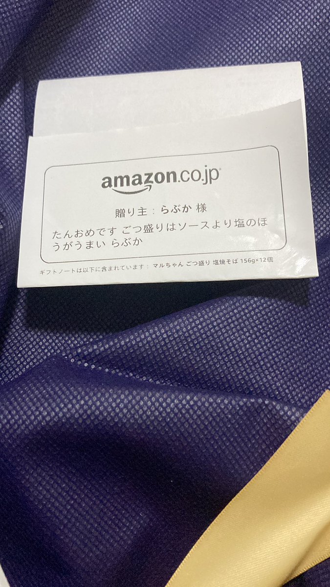 Purple and yellow instant ramen cups labeled as generous salt yakisoba from Amazon Japan, placed on a white envelope with Japanese text describing the product as five bags of thick noodles and sauce, viewed from above on a surface.