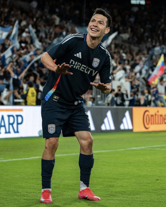 A male soccer player with short dark hair wearing a black Adidas San Diego FC jersey with DIRECTV sponsor logo, black shorts, black socks, and red cleats stands on a green soccer field in a stadium filled with spectators holding blue and white flags, gesturing with both hands extended forward in a celebratory pose, with stadium banners and sponsor ads visible in the background including ARP and Continental.