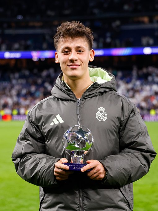 Jude Bellingham stands on a soccer field wearing a gray hooded jacket with Adidas logo and Real Madrid crest holding a shiny Champions League trophy smiling with stadium seating and blurred green pitch visible behind him.