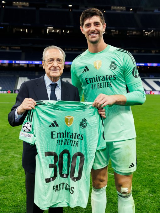 An elderly man with glasses wearing a dark suit and tie stands beside a tall athletic man in a green long-sleeved Real Madrid jersey numbered 25 on the field at night under stadium lights. Both hold a green Real Madrid jersey displaying the number 300 and Partidis on the back, along with the Champions League patch and sponsor logos like Emirates Fly Better. The setting is a soccer pitch with grass and stadium seating in the background.