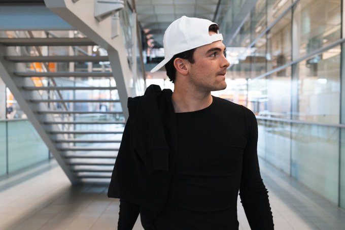 A young man with short hair wearing a white cap backwards and a black long-sleeve shirt stands in a modern indoor space with glass walls and metal stairs in the background, looking to the side with a slight smile while holding a black jacket over his shoulder.