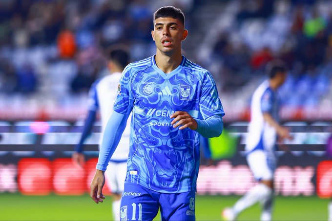 Player wearing blue Tigres jersey with number 11 and long sleeves stands on soccer field during match. Blue and white stadium crowd visible in background. Sponsor logos on jersey including Cemex. Player has dark hair and focused expression while walking.