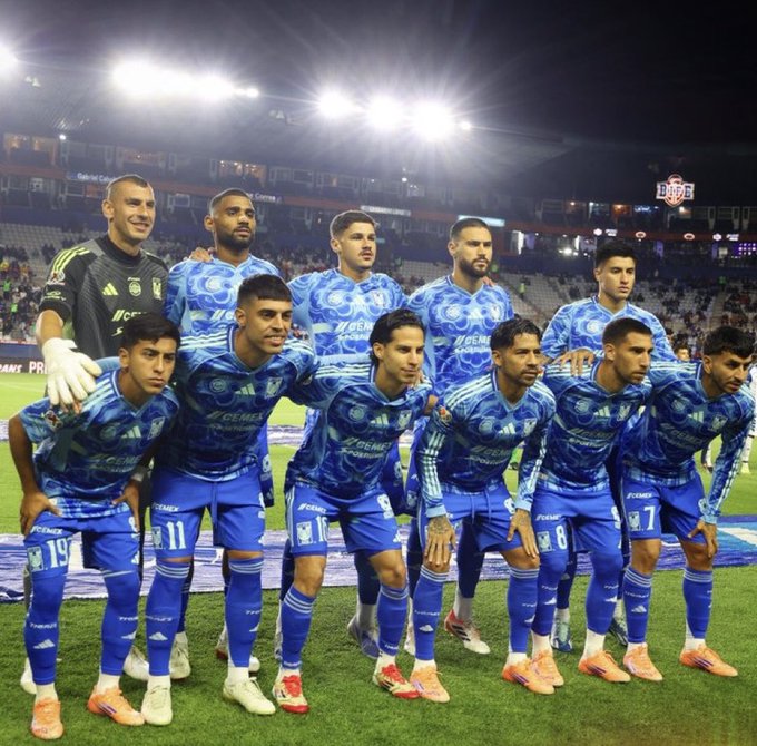 Group of eleven male soccer players in blue Tigres uniforms with white accents and numbers like 11, 8, 7, standing arm-in-arm on a green field at night under bright stadium lights, with a goalkeeper in green jersey and gloves positioned separately, orange-shoed feet visible, blurred crowd and scoreboard in background.