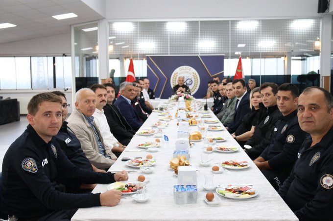 First image shows a long conference table with multiple men and women in dark blue police uniforms and civilian attire seated around it, engaged in eating breakfast including plates of vegetables, bread, cheese, and tea bottles, with Turkish flags and a blue emblem backdrop in a brightly lit room. Second image depicts a similar group setting with more officers and civilians at the table, flowers in the center, Düzce Polis written on the backdrop, and flags on sides. Third image features a smaller group including uniformed female and male officers at a round table with breakfast items like tea pots, cups, and food platters, microphones present, and Düzce emblem visible.