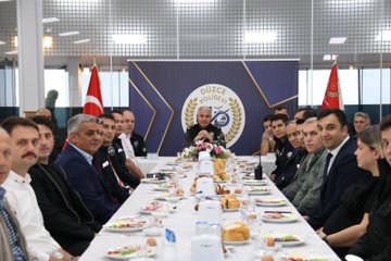 First image shows a long conference table with multiple men and women in dark blue police uniforms and civilian attire seated around it, engaged in eating breakfast including plates of vegetables, bread, cheese, and tea bottles, with Turkish flags and a blue emblem backdrop in a brightly lit room. Second image depicts a similar group setting with more officers and civilians at the table, flowers in the center, Düzce Polis written on the backdrop, and flags on sides. Third image features a smaller group including uniformed female and male officers at a round table with breakfast items like tea pots, cups, and food platters, microphones present, and Düzce emblem visible.