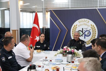 First image shows a long conference table with multiple men and women in dark blue police uniforms and civilian attire seated around it, engaged in eating breakfast including plates of vegetables, bread, cheese, and tea bottles, with Turkish flags and a blue emblem backdrop in a brightly lit room. Second image depicts a similar group setting with more officers and civilians at the table, flowers in the center, Düzce Polis written on the backdrop, and flags on sides. Third image features a smaller group including uniformed female and male officers at a round table with breakfast items like tea pots, cups, and food platters, microphones present, and Düzce emblem visible.
