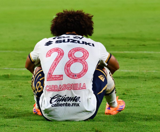 A soccer player with curly hair wearing a white jersey with number 28 in red on the back and name Carrasquilla above it kneels on green grass field the jersey has sponsor logos including Suzuki and Caliente.mx on the bottom the player wears navy shorts with gold accents orange cleats and navy socks with gold stripes the background shows part of the soccer field