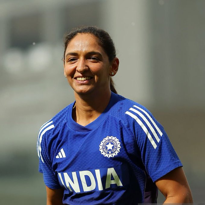 Harmanpreet Kaur stands on a cricket field wearing a blue India national team jersey with the BCCI emblem and India text on the front, smiling at the camera with her hair tied back and small earrings visible, background shows blurred stadium seating and lighting.